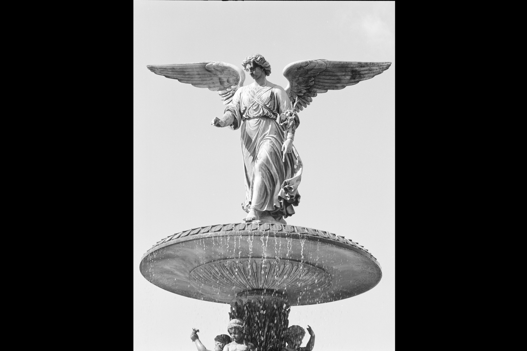 Bethesda Fountain: A black-and-white photograph shows a winged angel statue standing atop a large fountain, arms gently extended as water cascades below against a bright, open sky.
